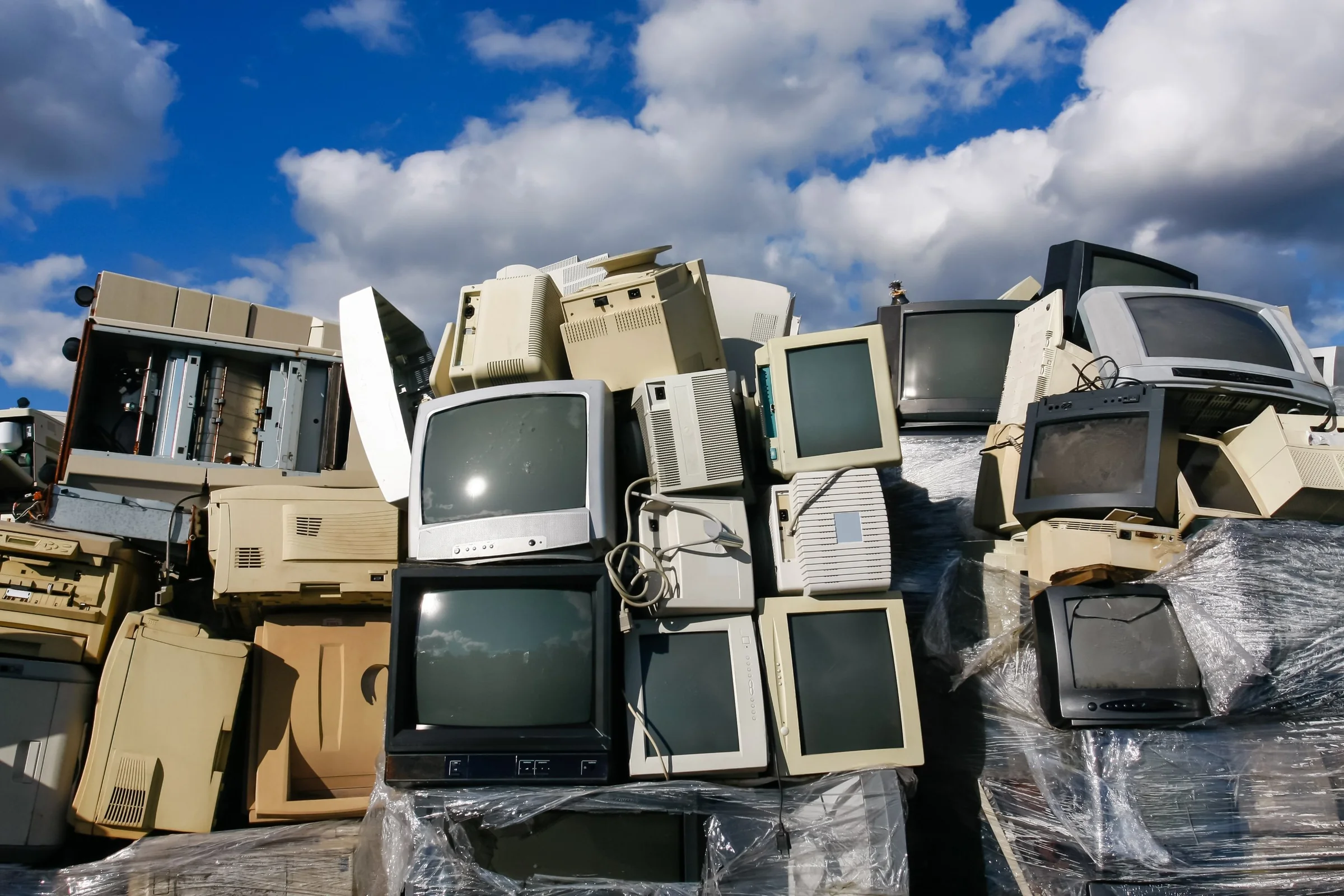 Old tube TVs, beige computer monitors and printers stacked up outdoors under a bright blue sky