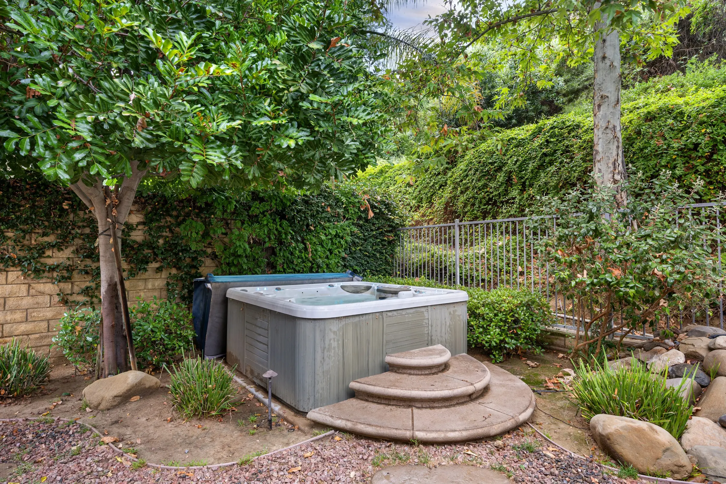 An older hot tub with a faded gray cabinet and stone steps, sitting in a shady corner of a backyard