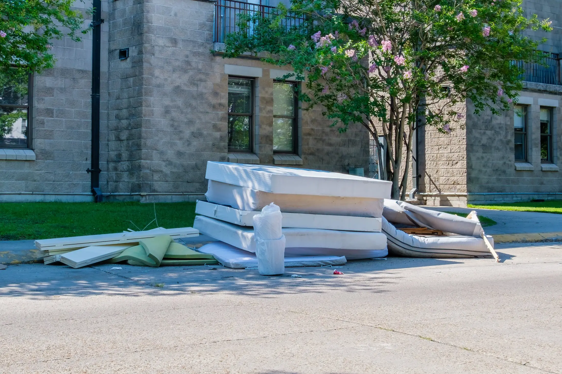 Worn white mattresses and a box spring stacked at the curb beside a stone building