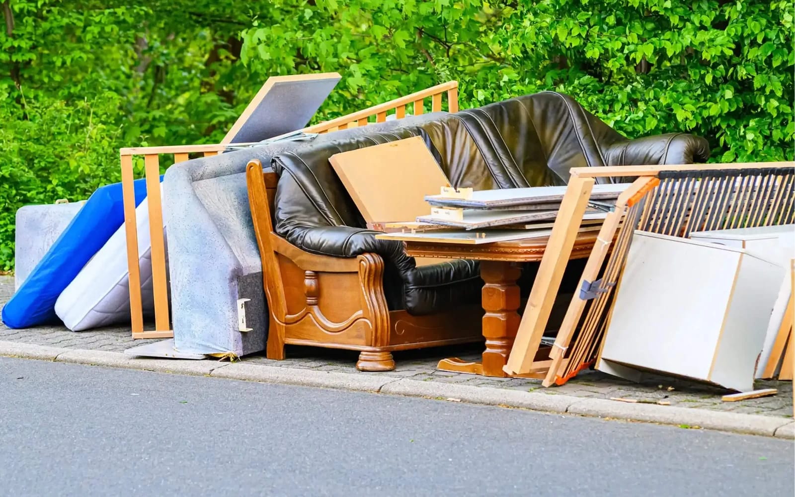 Old furniture piled at the curb, including a black leather sofa, a carved wooden armchair, mattresses, bed frames and a table