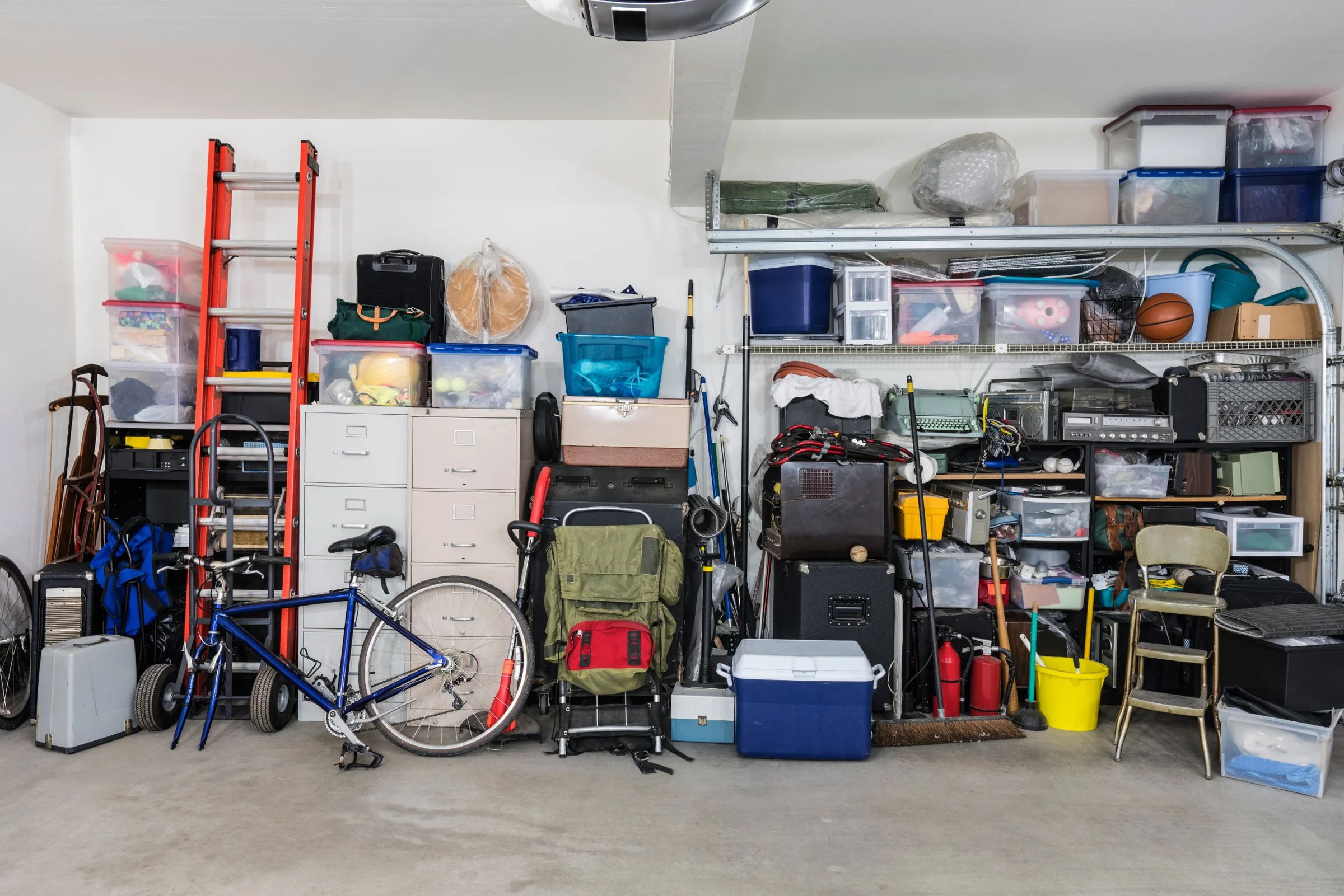 A packed garage wall with storage bins up to the ceiling, a ladder, a bicycle, filing cabinets and a cooler
